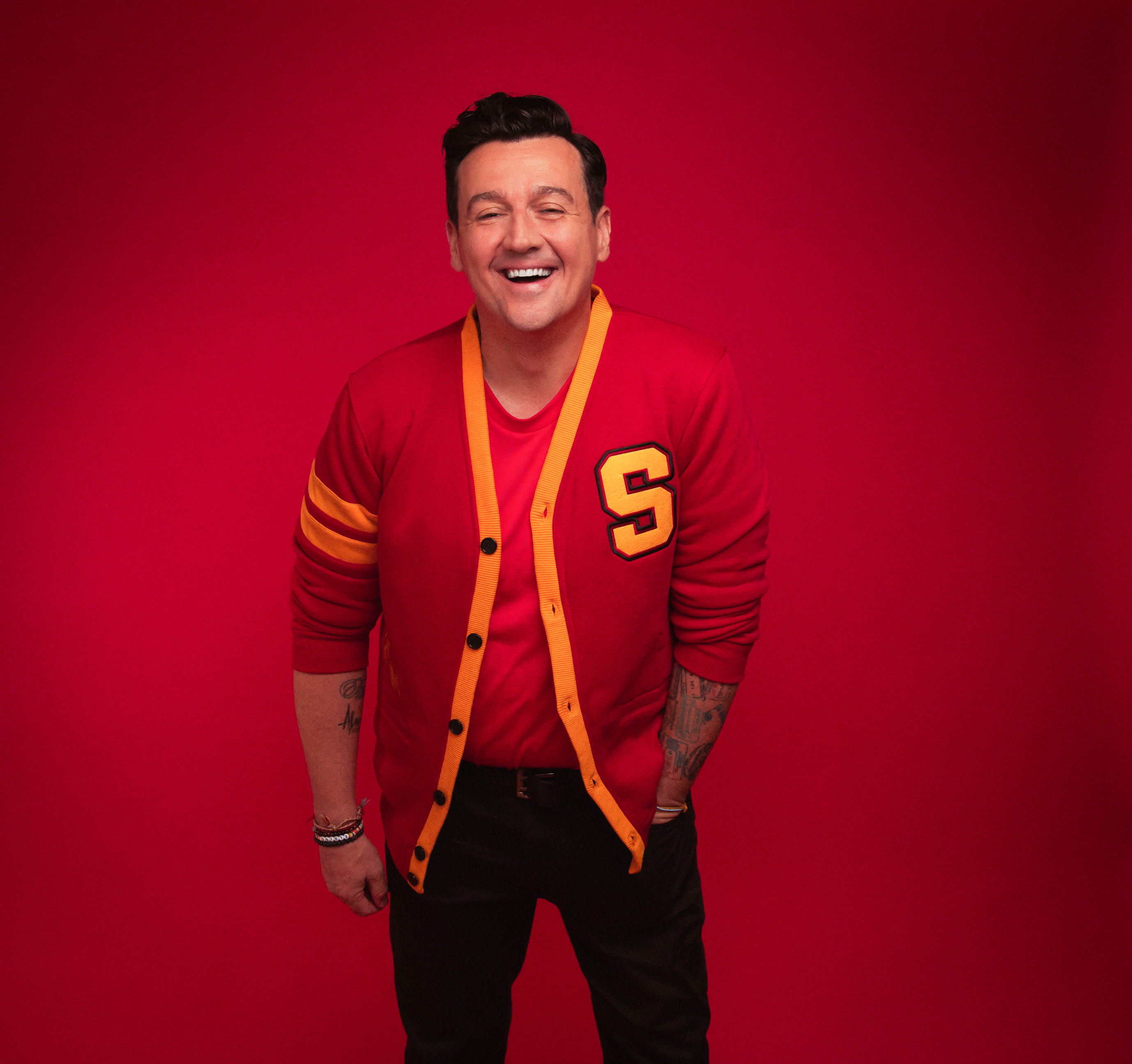 Sam Moran laughing with his hands in his pockets against a red backdrop