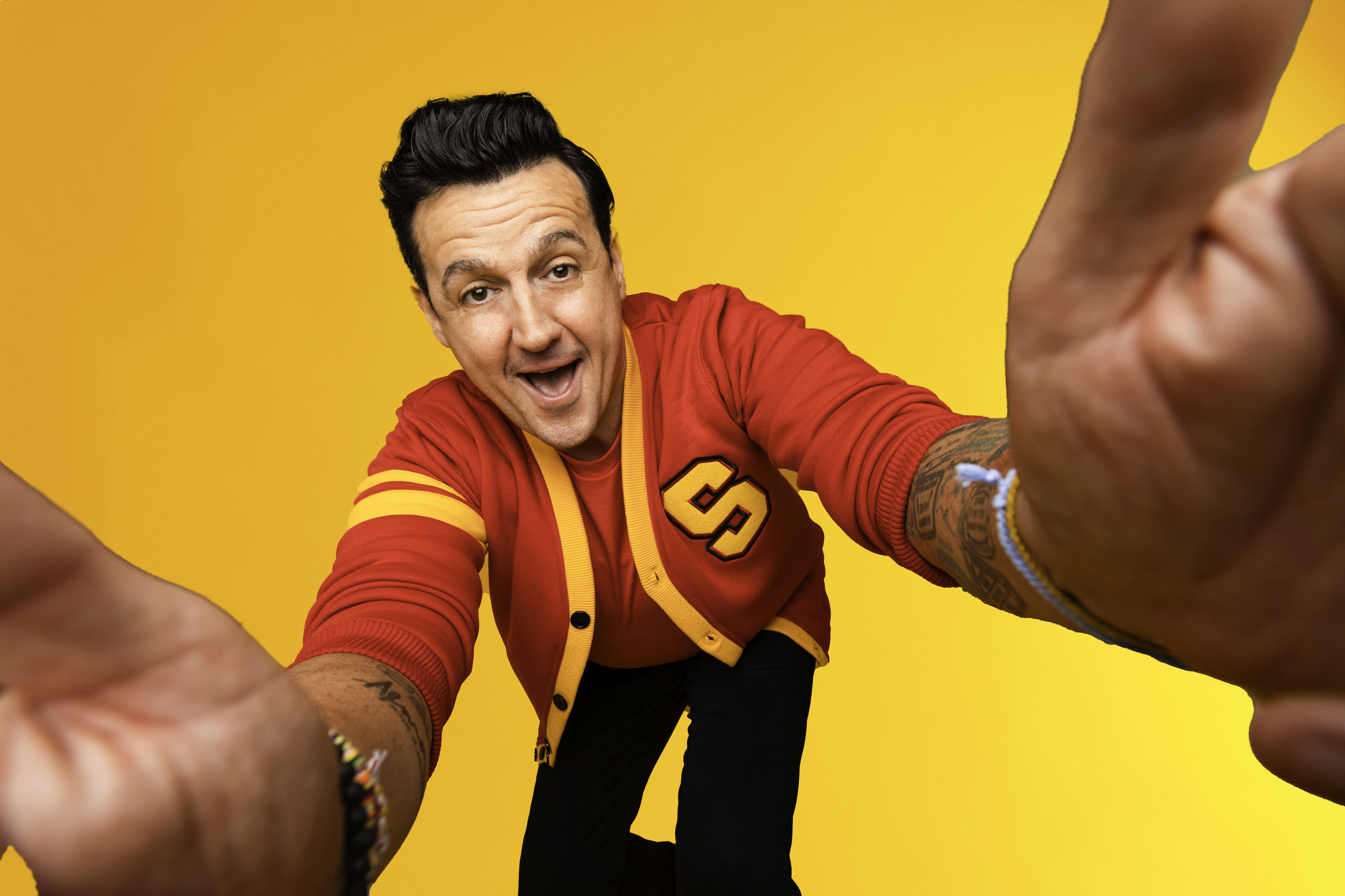 Sam Moran in a red varsity cardigan reaching both hands towards the camera against a yellow backdrop