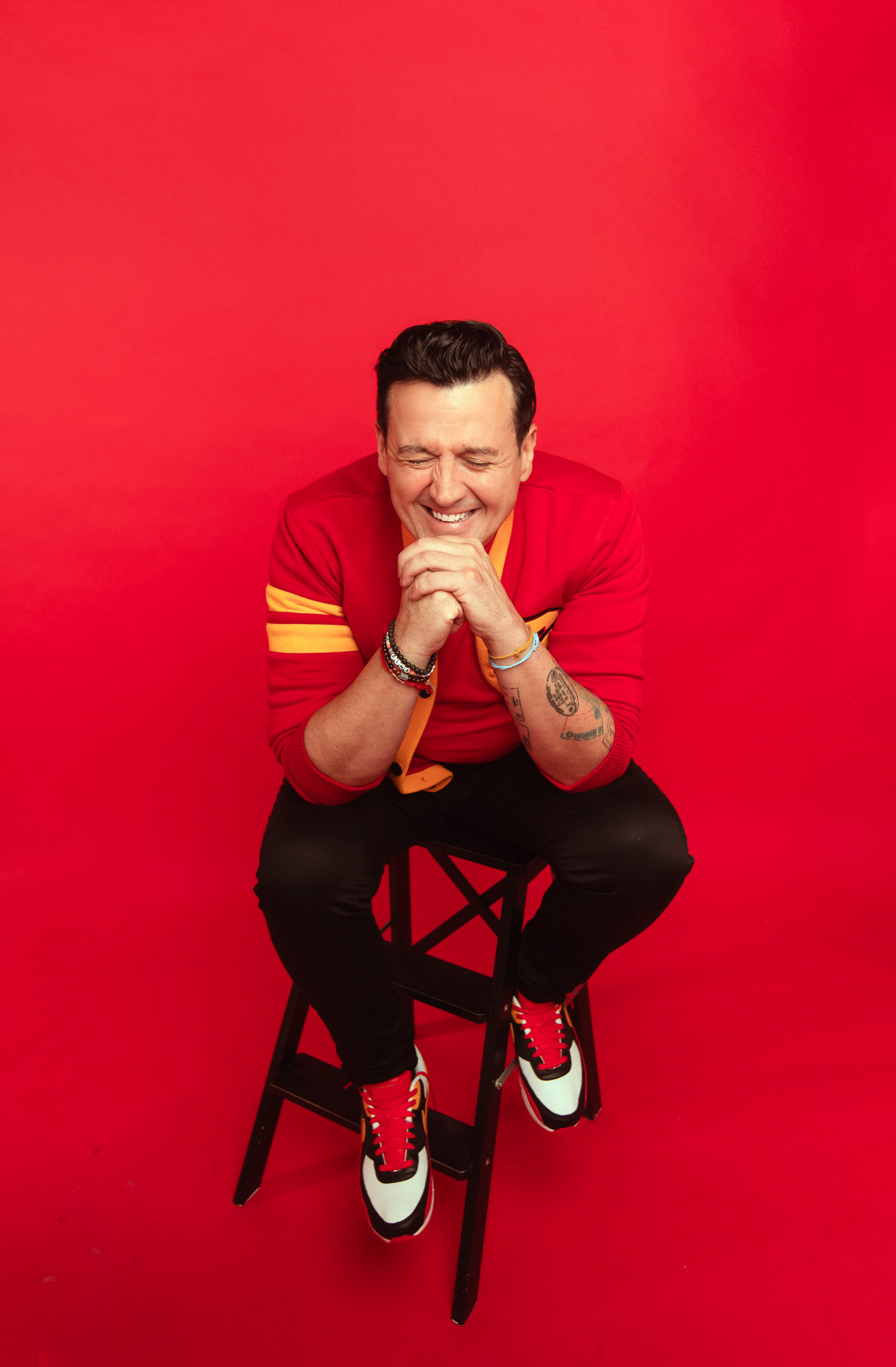 Sam Moran sitting on a stool with hands clasped, laughing, against a red backdrop