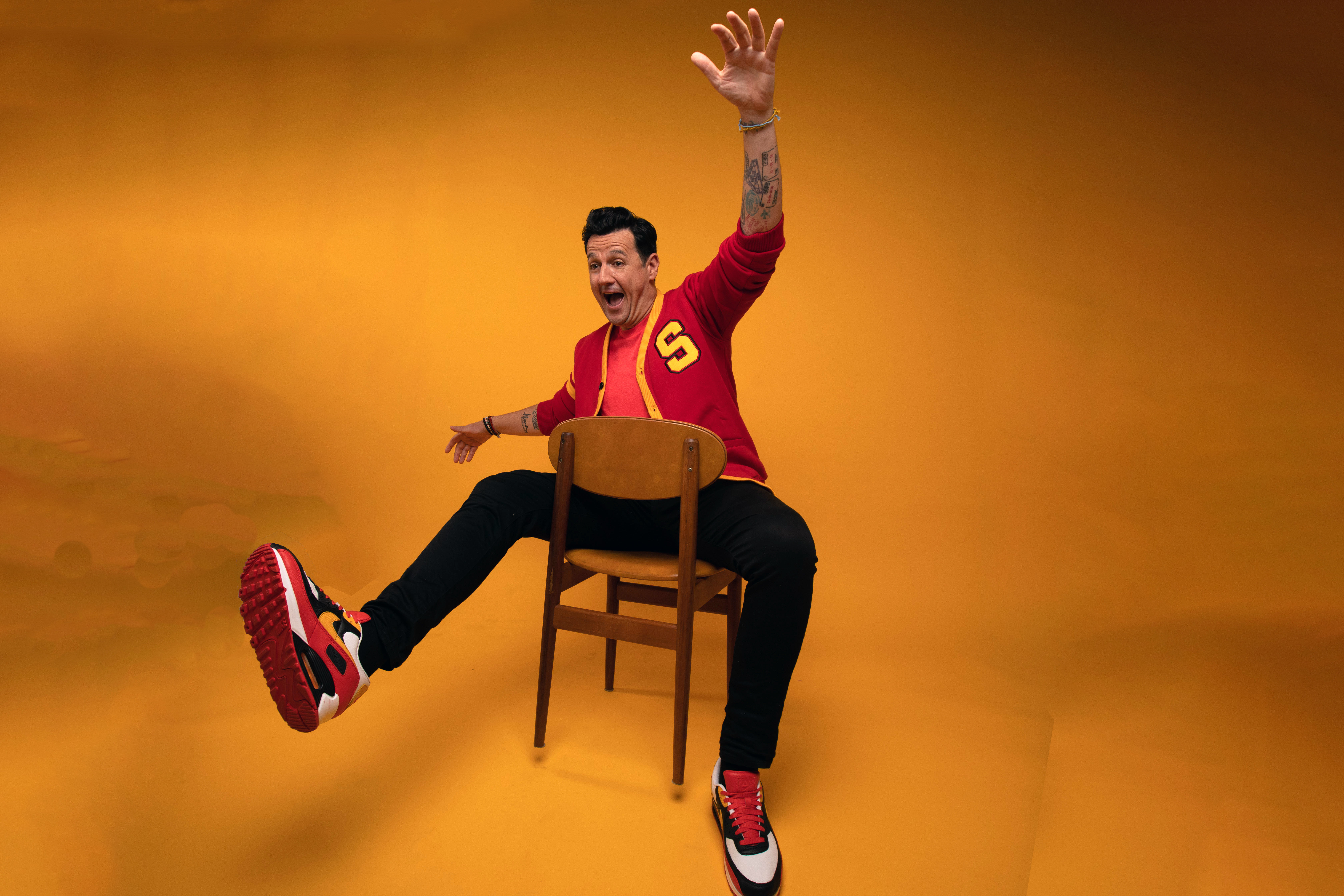 Sam Moran sitting sideways on a wooden chair with one leg kicked out and an arm raised, against an orange backdrop